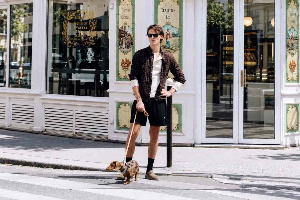 Homme portant une chemise à carreaux et un short noir, promenant son chien dans une rue parisienne.