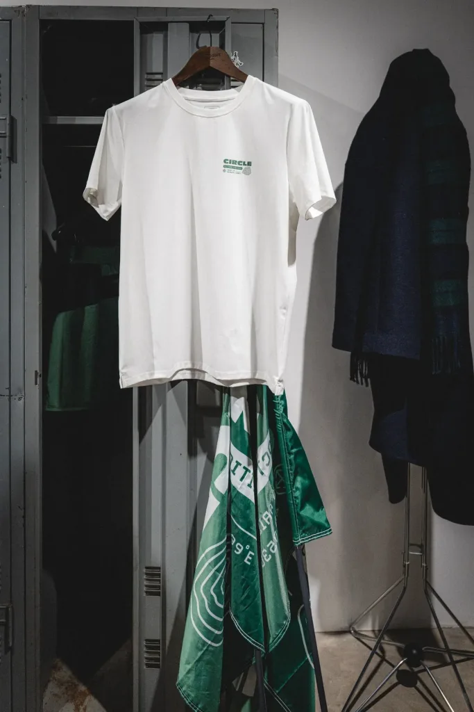 T-shirt écru Circle x Octobre Éditions suspendu dans un vestiaire, accompagné d'un textile vert imprimé.
