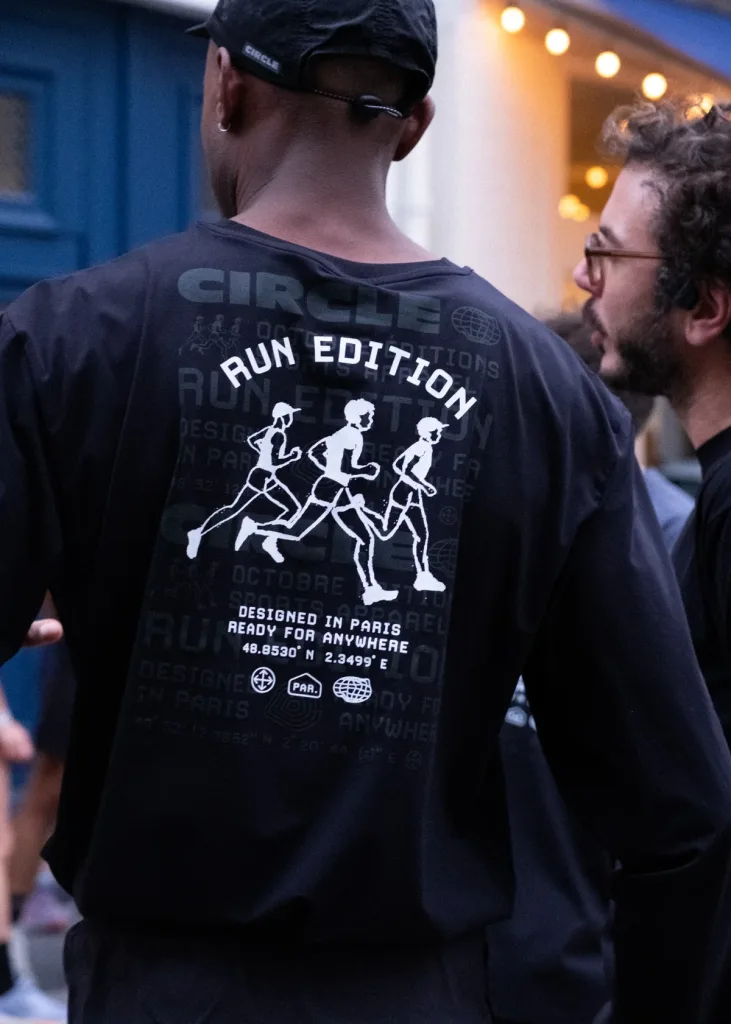 Vue de dos d'un t-shirt noir Circle x Octobre Éditions avec un imprimé blanc représentant trois coureurs.